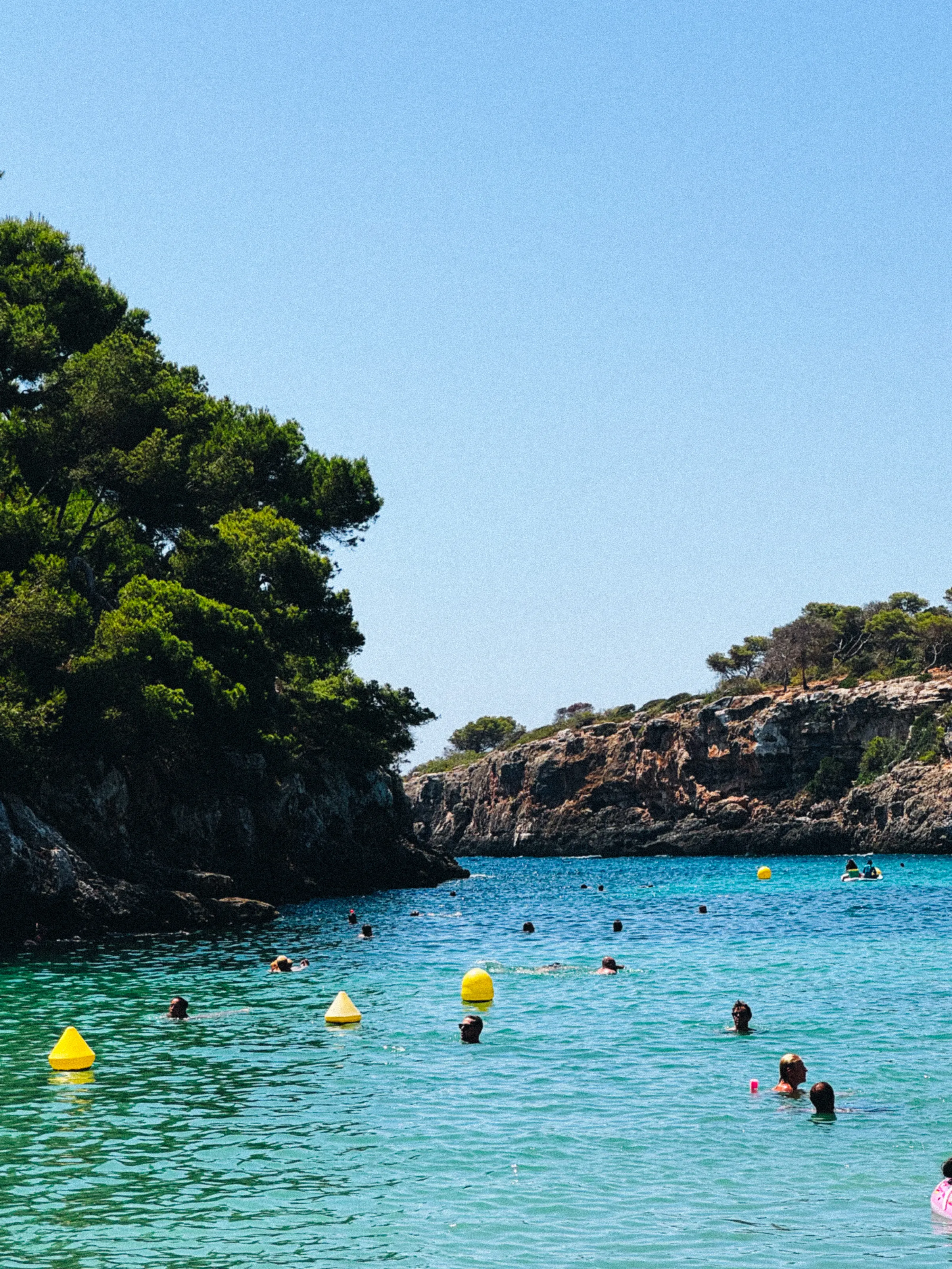 A rocky cove with swimmers using pool noodles, seen from a shaded ledge above the water