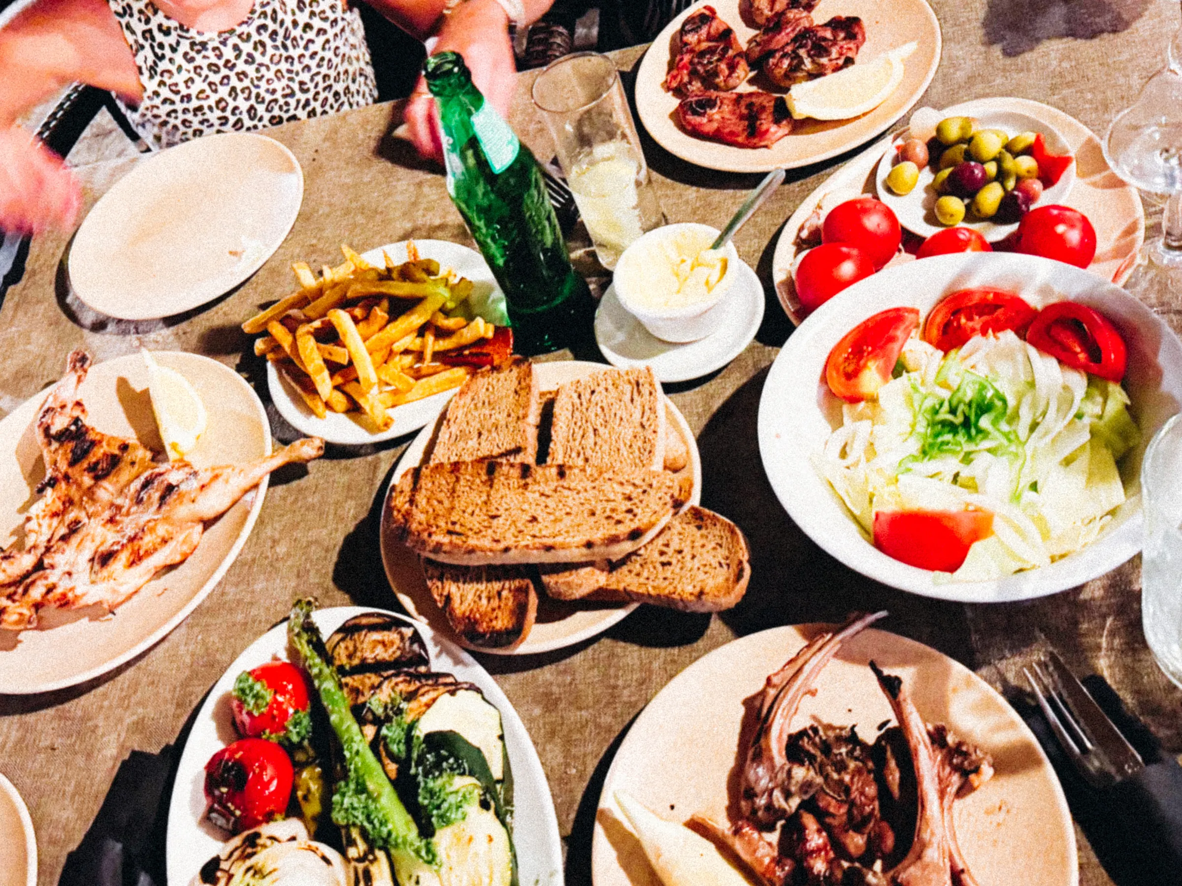 A shared table crowded with grilled meats, bread, fries, salad, tomatoes, and olives