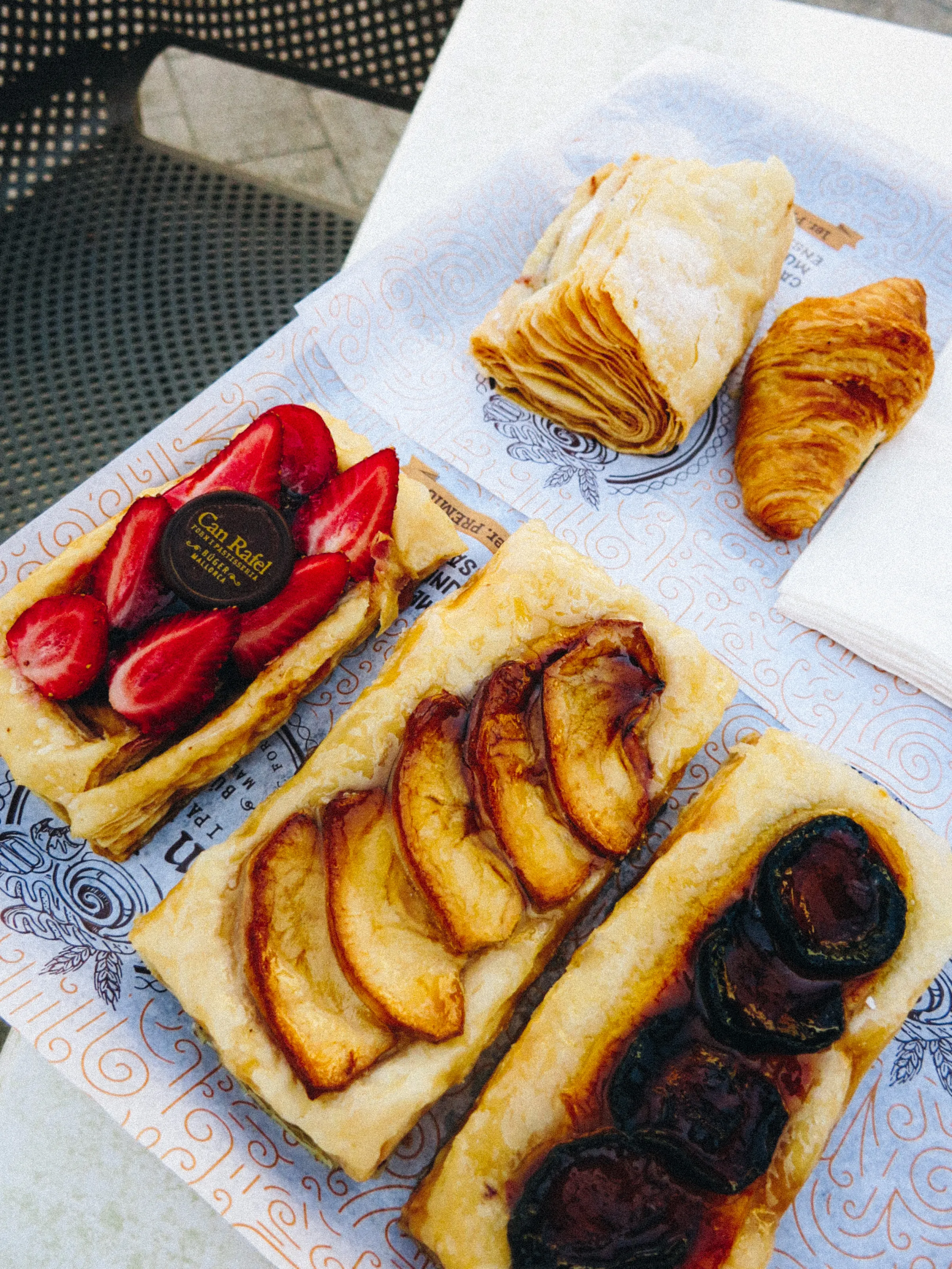 Pastries on a café table: a strawberry tart, an apple tart, a plum tart, and a croissant