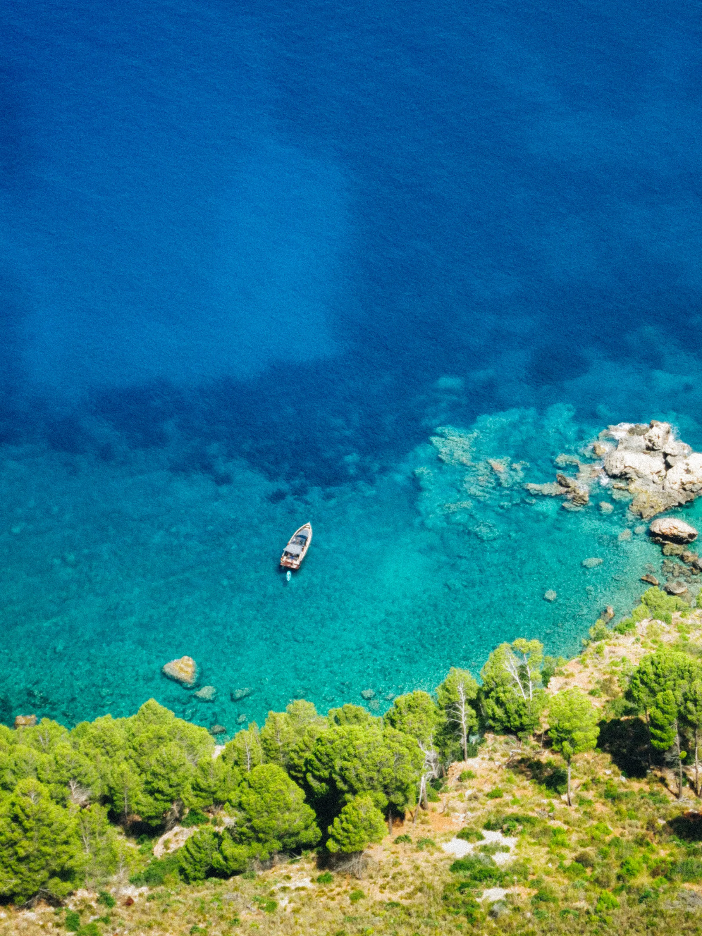 An aerial view of a small boat anchored in a turquoise cove ringed by pine trees