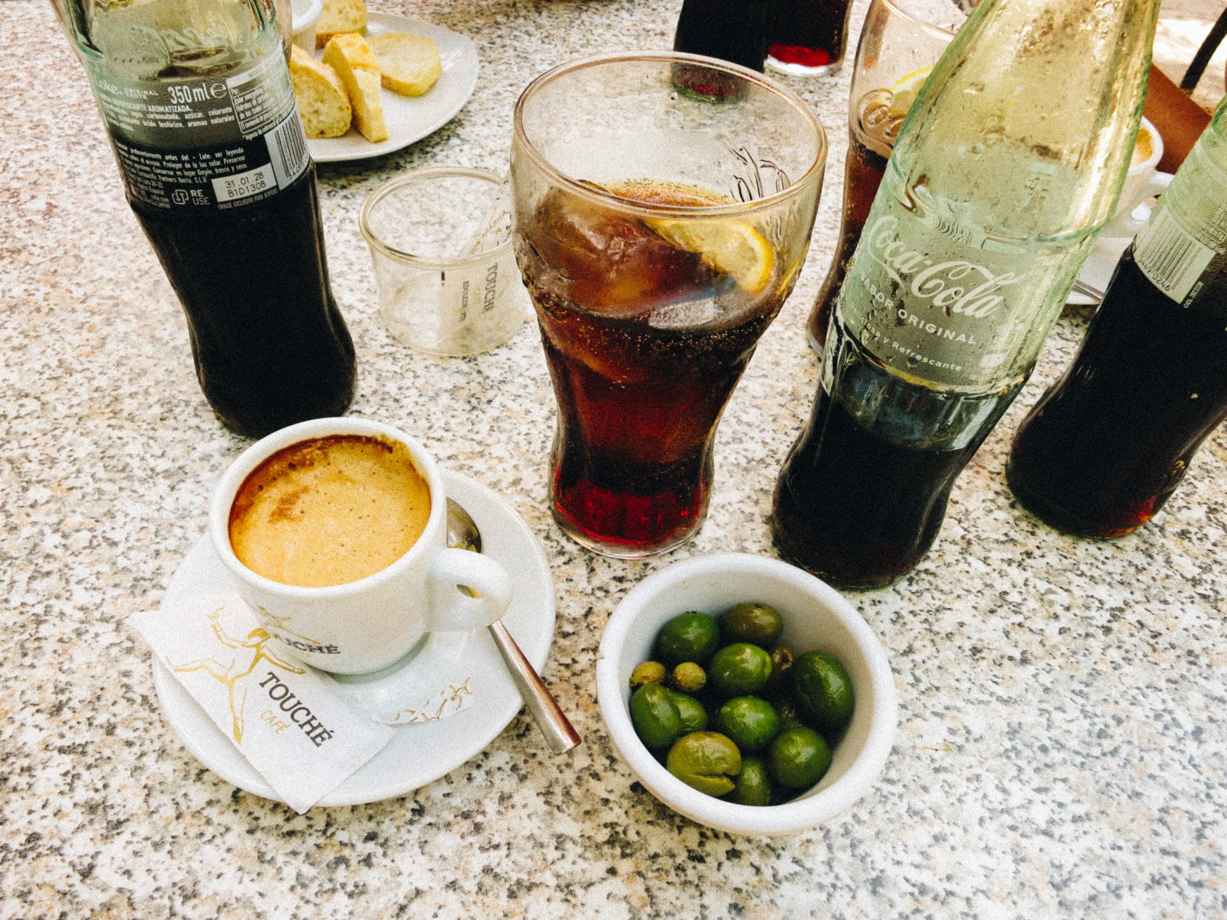 A café table with an espresso, a glass of cola with lemon, and a bowl of green olives