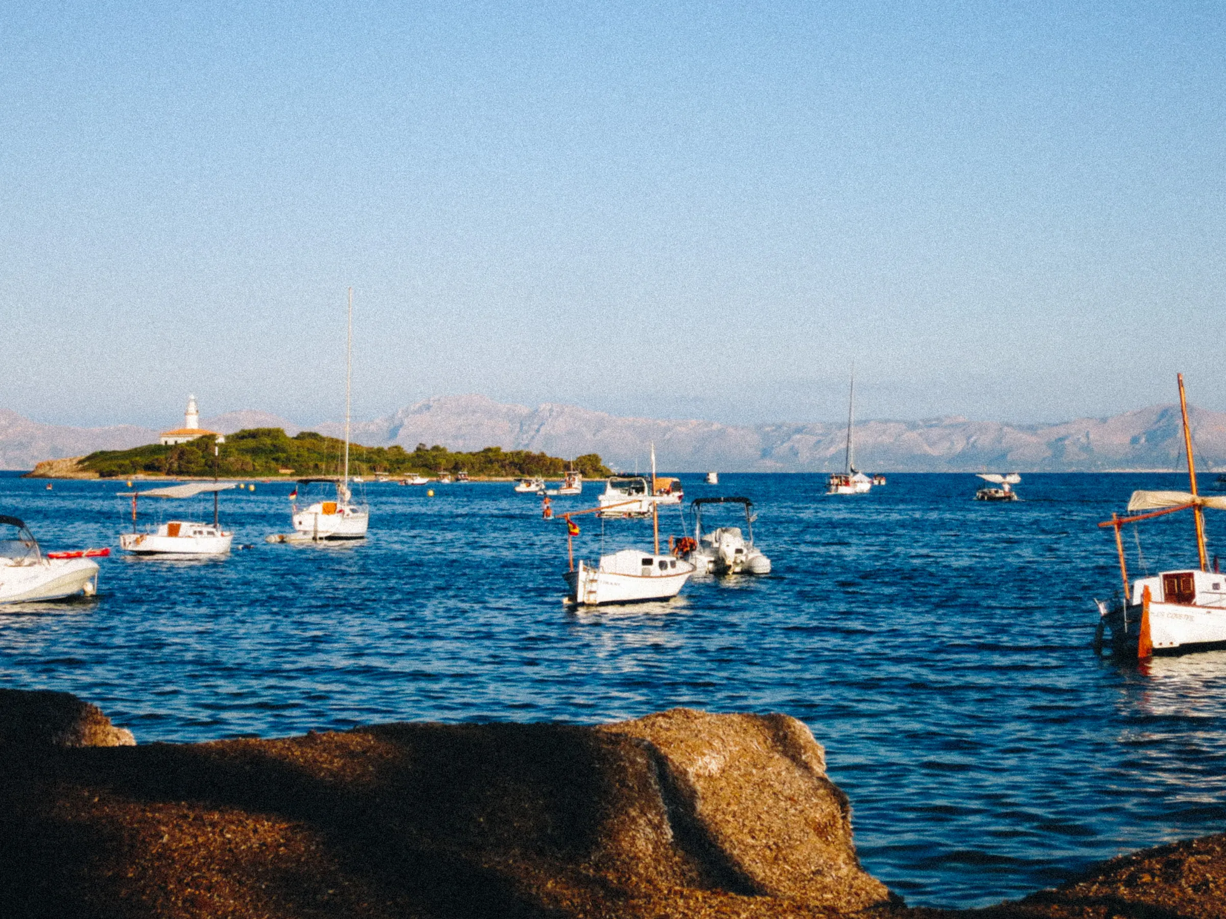 A small harbor with boats moored near a lighthouse on a rocky island, mountains in the distance