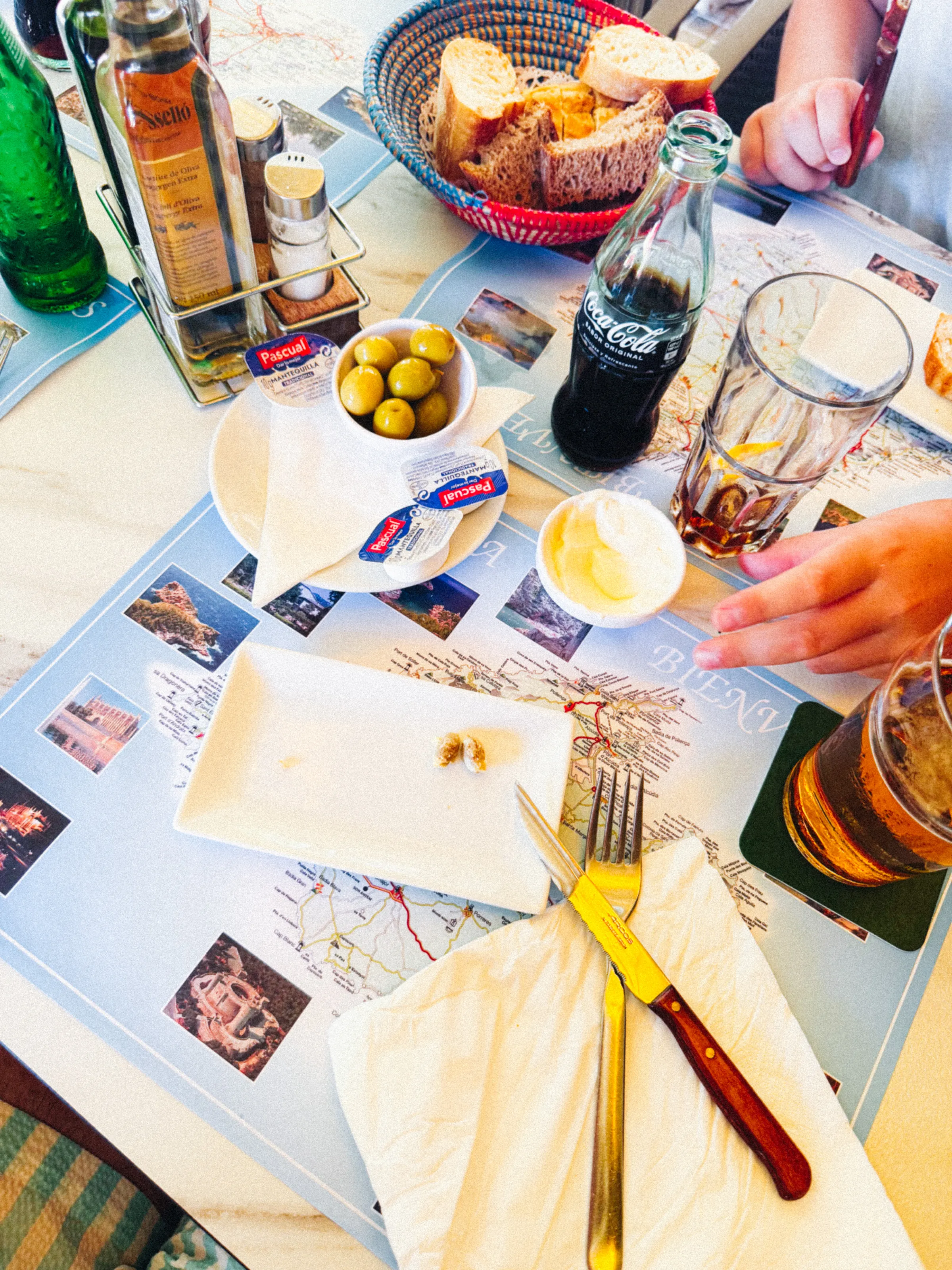 A café table with a bread basket, a Coca-Cola bottle, a bowl of olives, butter, and an empty shell plate