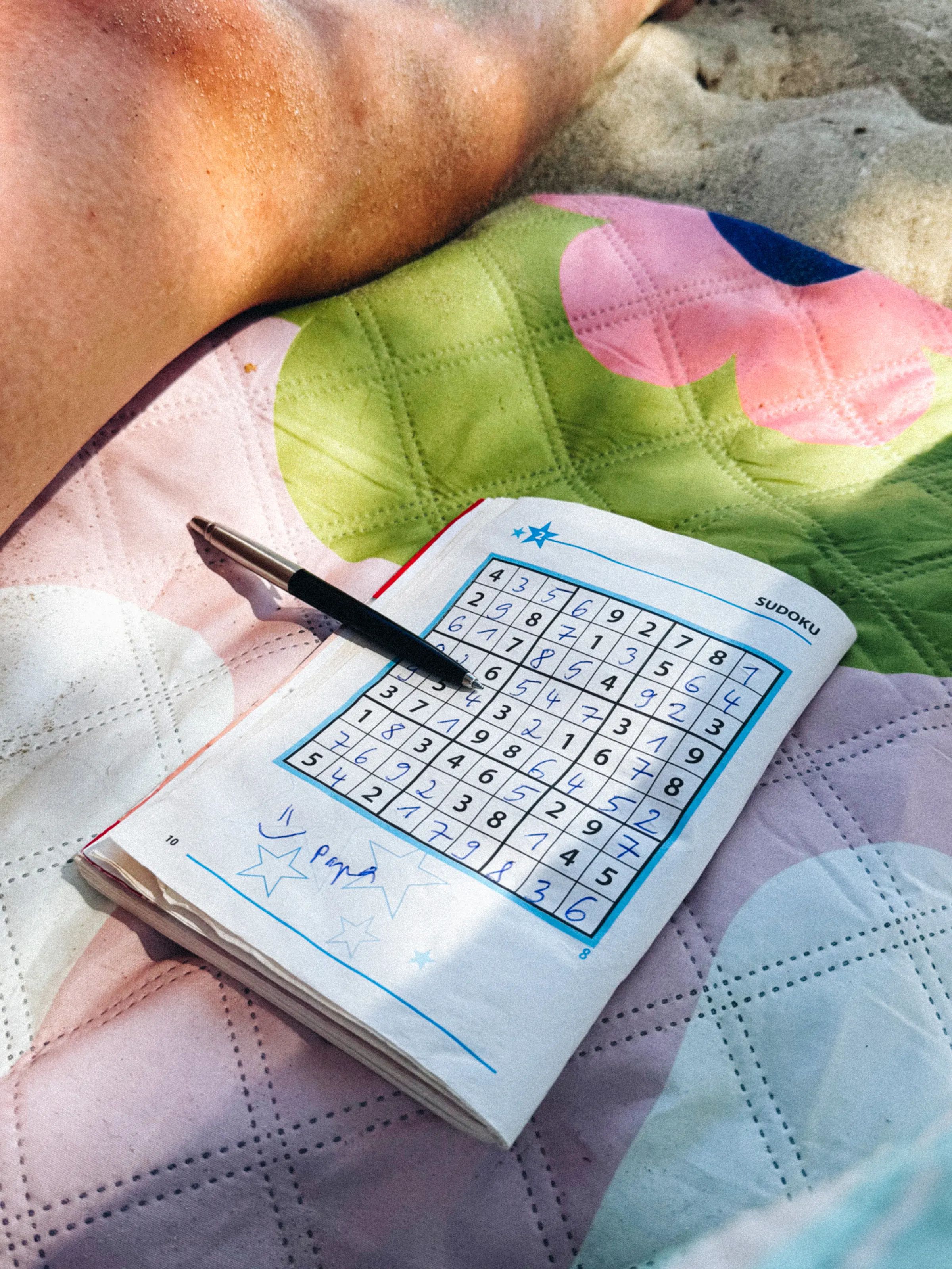A sudoku booklet and pen resting on a floral beach towel next to a sunburned arm