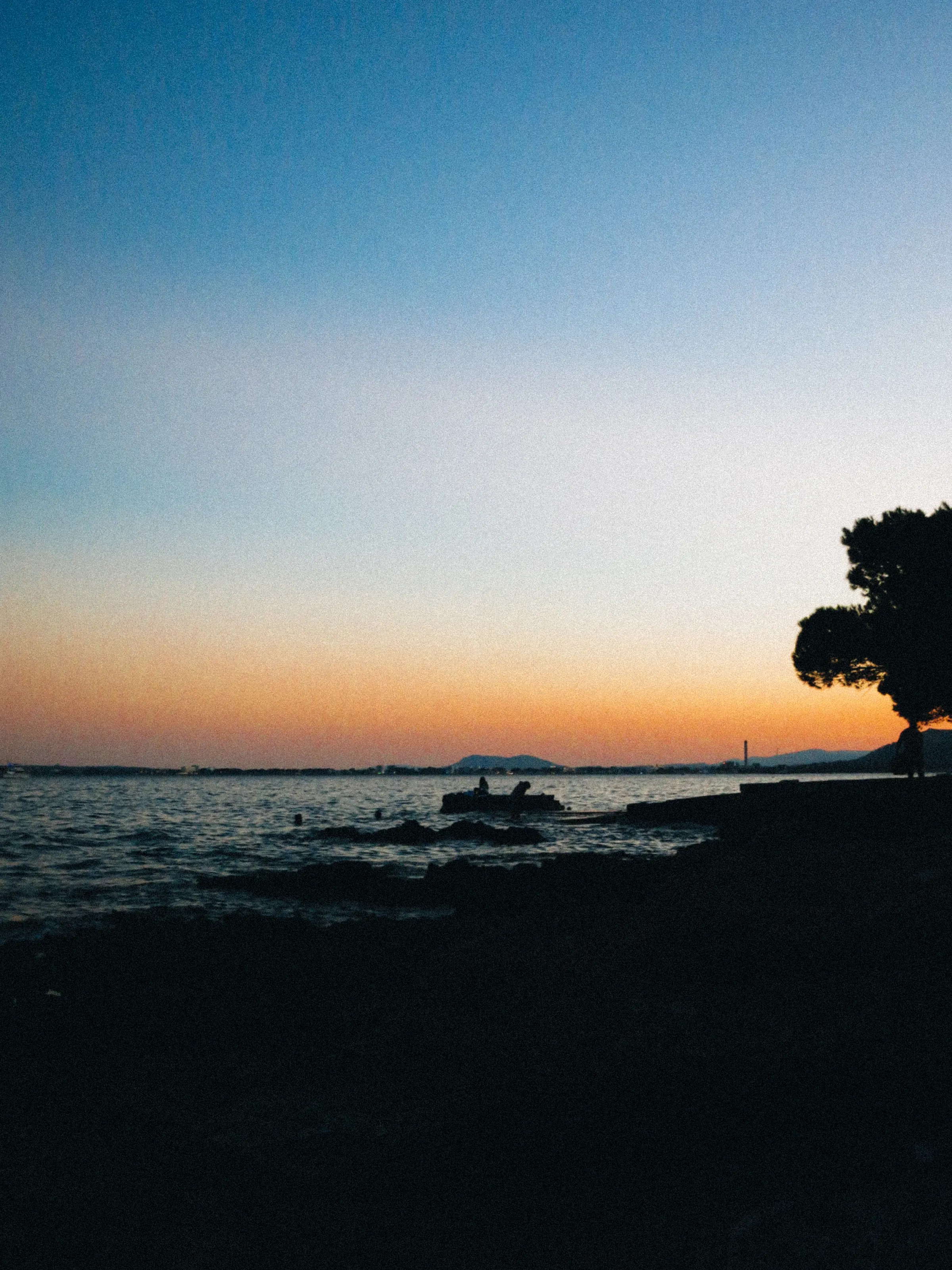 A silhouetted boat and swimmers at sunset, the sky fading from orange to blue
