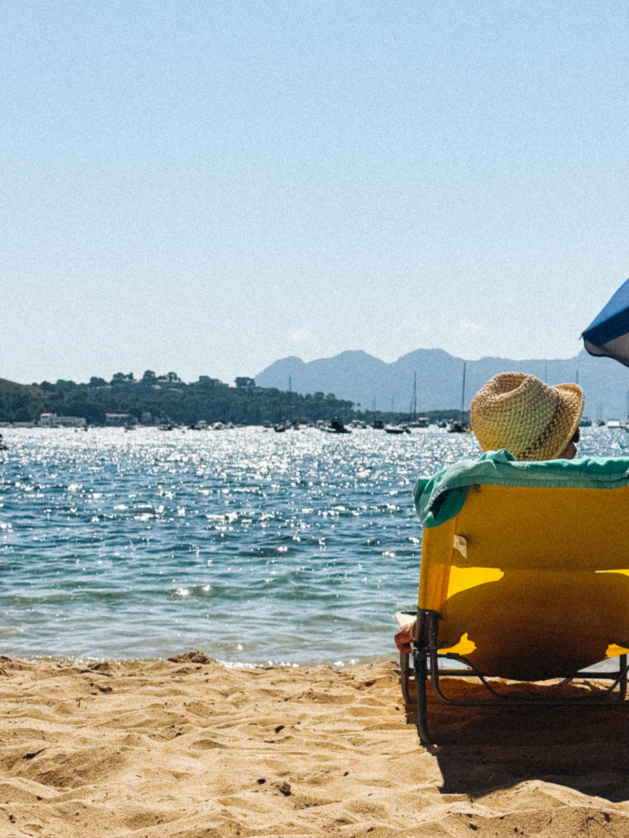 A person in a straw hat sitting in a yellow beach chair, facing a sparkling bay with hills across the water