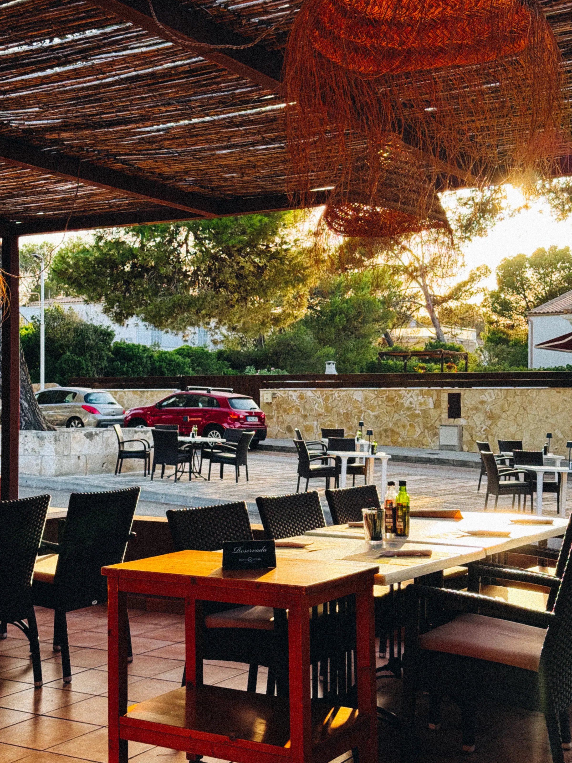 An outdoor restaurant terrace with a reed roof, tables set for dinner, in warm evening light