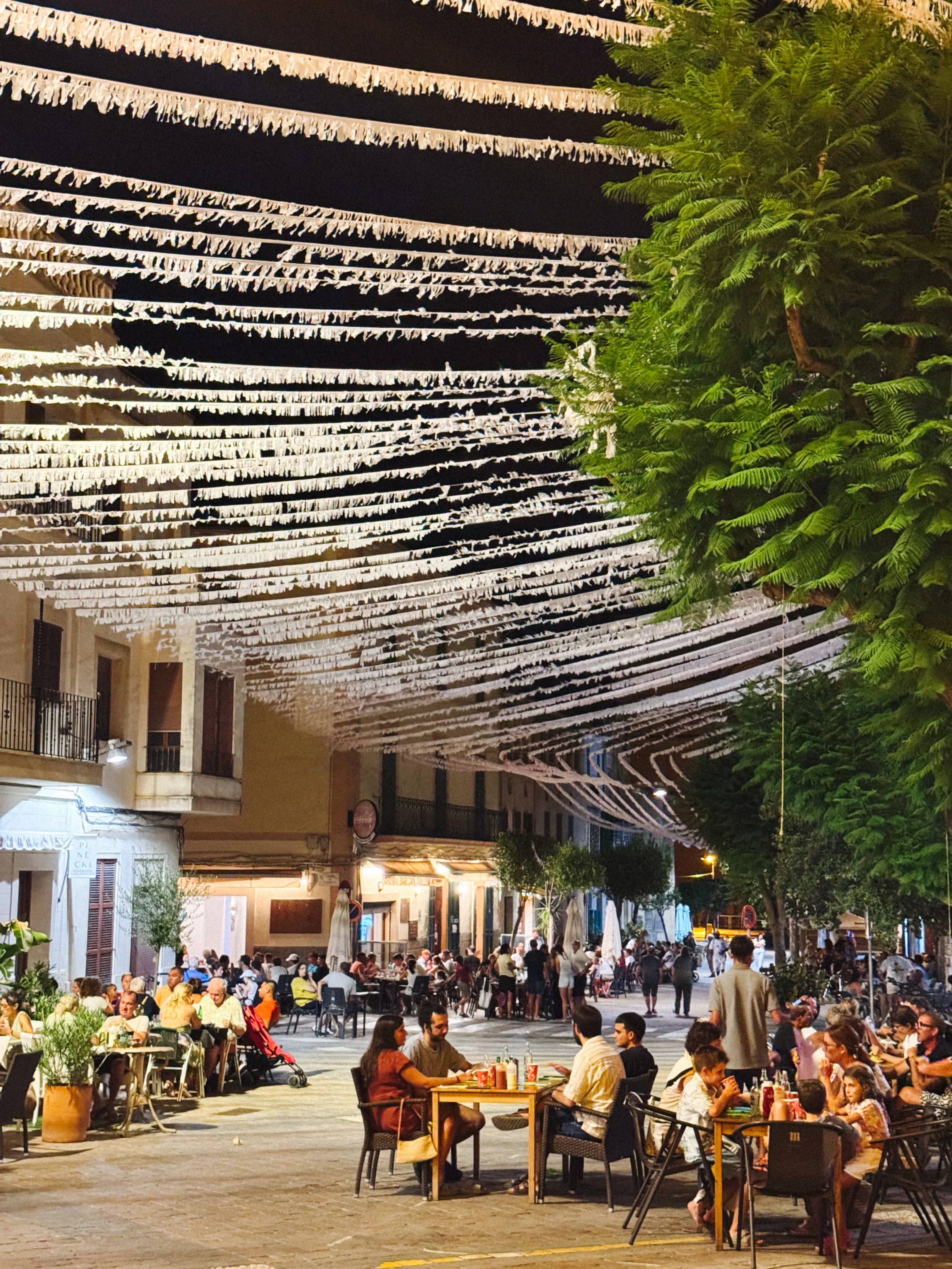 A town square at night strung with rows of white paper streamers overhead, people dining at outdoor tables